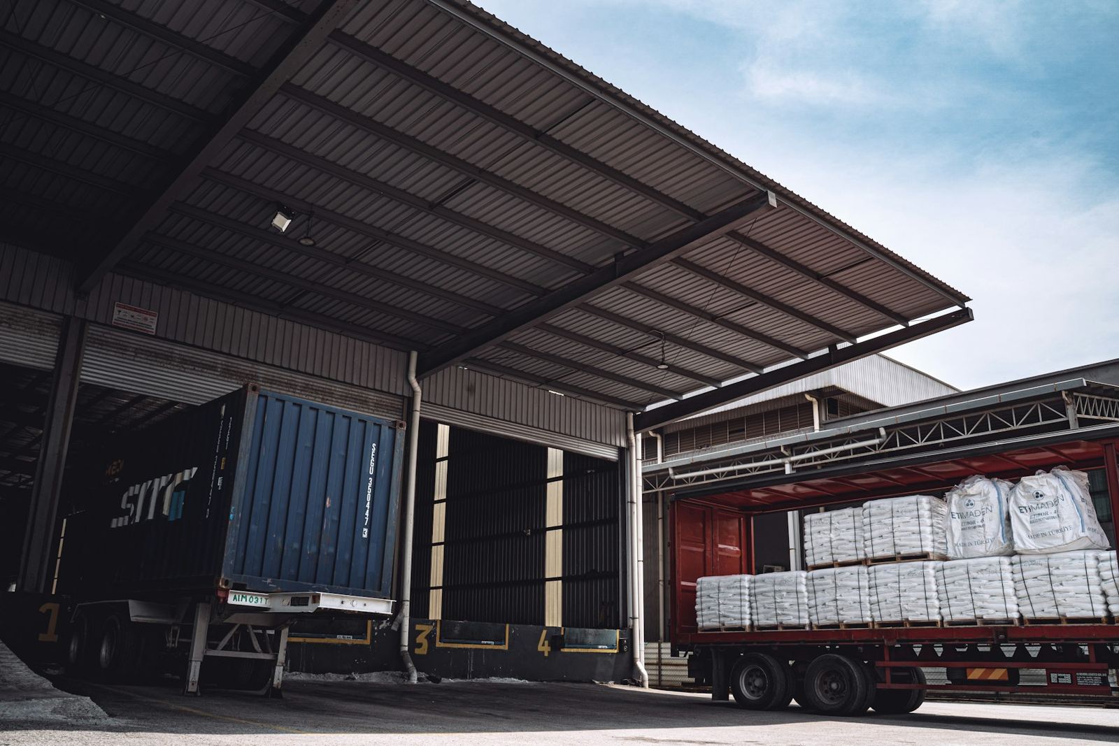 A container truck backed into a covered loading dock, with a second truck being loaded with white sacks